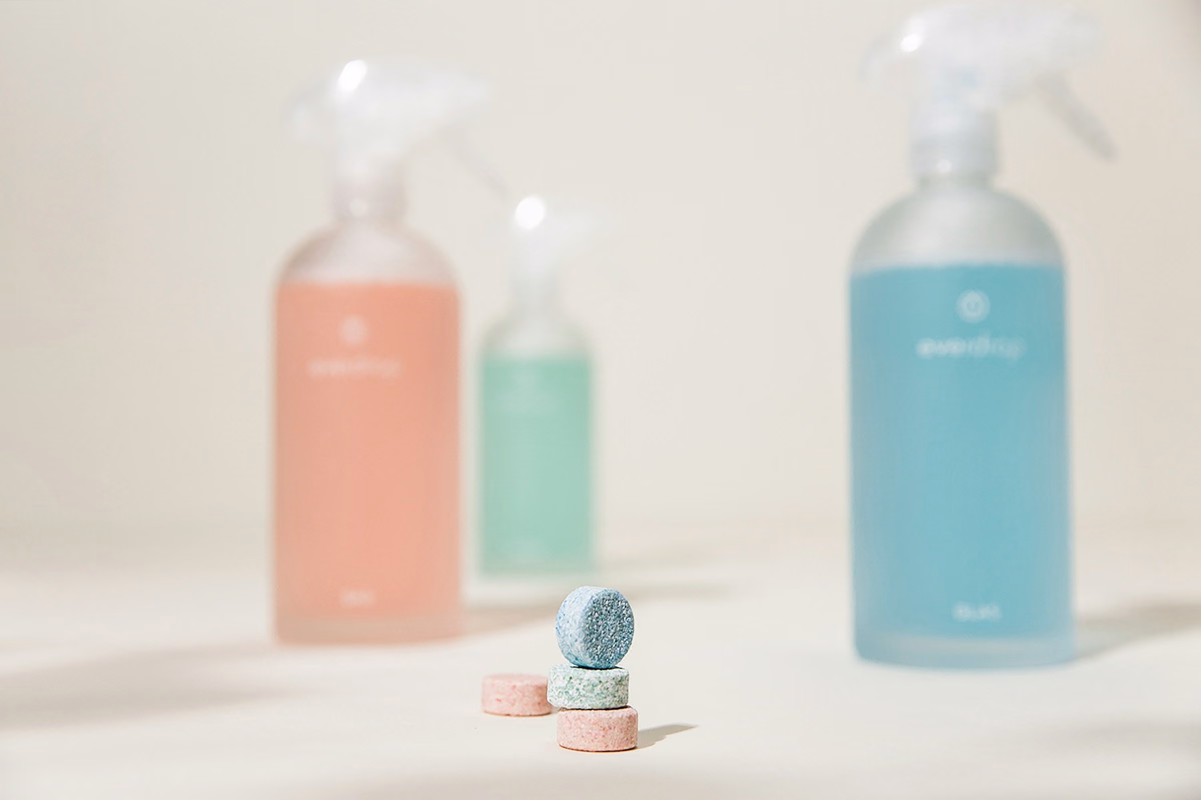Stack of three colorful cleaning tablets in front of three spray bottles with pastel-colored liquids.