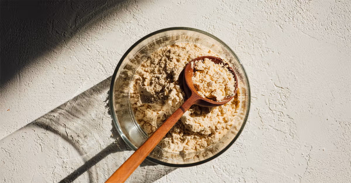 Glass bowl with light beige powder and a wooden spoon filled with the powder on a textured surface.