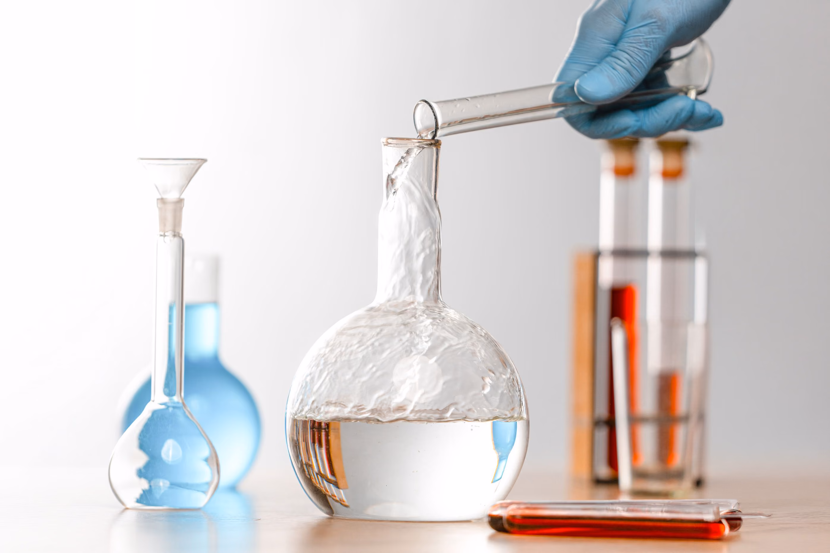 Hand in blue glove pouring clear liquid from test tube into round bottom flask in lab setting.