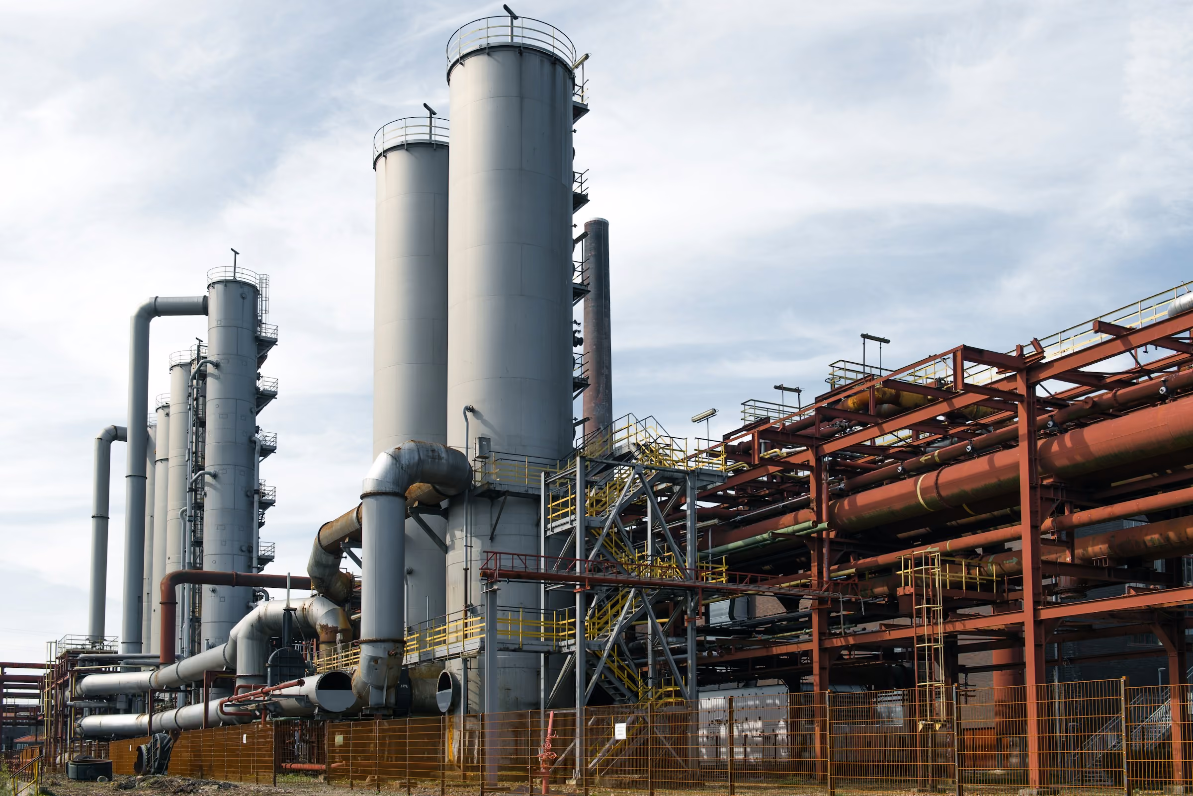 Large industrial refinery complex with tall cylindrical tanks and extensive piping under a cloudy sky.